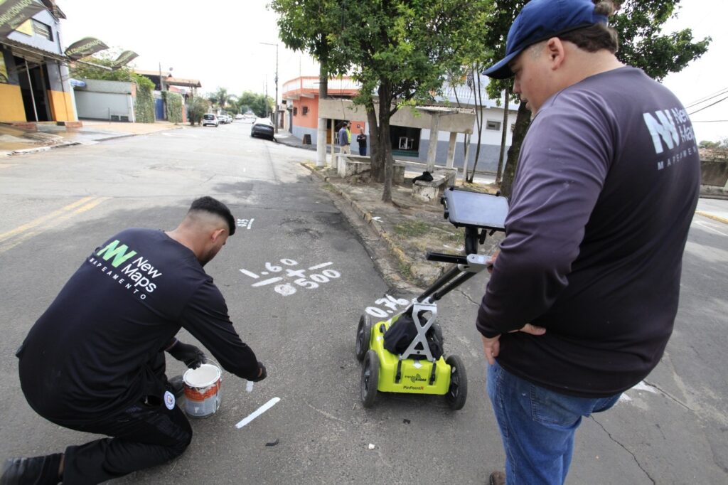 A imagem mostra uma cena ao ar livre em uma rua pavimentada, onde dois homens estão realizando um mapeamento utilizando um equipamento de Radar de Penetração no Solo (GPR). Ambos vestem camisas escuras de manga longa com o logotipo "New Maps MAPEAMENTO". Um dos homens está agachado no chão, pintando marcas brancas na superfície do asfalto com um pincel, ao lado de uma lata de tinta. O outro homem está em pé, observando um tablet conectado ao equipamento GPR, que é amarelo com rodas pretas e identificado como "PinPointR". No chão, há diversas marcações numéricas pintadas, como "1.60", "0.500" e "0.35", provavelmente indicando medições ou profundidades relacionadas ao mapeamento. Ao fundo, vê-se uma rua residencial com algumas árvores, casas e poucas pessoas. A cena parece estar sendo realizada durante o dia, com iluminação natural e céu nublado.