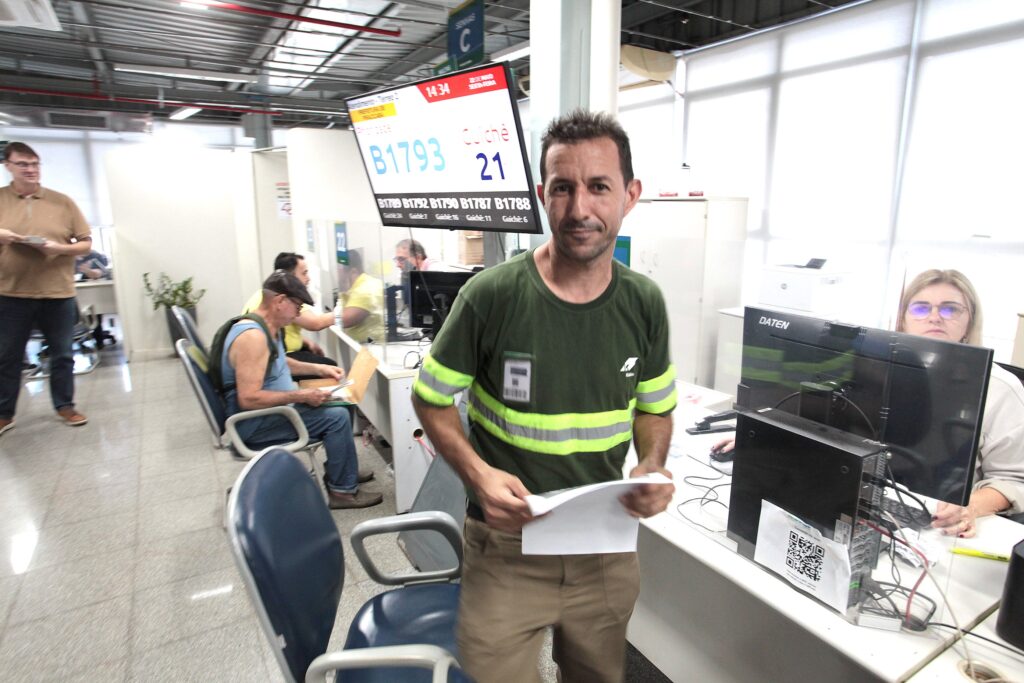A imagem mostra o interior de um ambiente de atendimento público ou administrativo, possivelmente uma agência governamental ou de serviços. * Em primeiro plano, um homem de meia-idade olha diretamente para a câmera, vestindo uma camiseta verde com faixas refletivas horizontais, típica de uniforme de trabalho, e calças cáqui. Ele segura um papel branco nas mãos. * Ao fundo, um painel eletrônico exibe o número de atendimento B1793, indicando o guichê 21, além da data e hora: 14:34, 22 de maio, terça-feira. * O espaço é organizado com baias de atendimento. À direita, uma mulher com óculos trabalha em um computador da marca DATEN. À esquerda, há outros clientes sentados aguardando atendimento, incluindo um homem de boné e camiseta azul sem mangas, e outro homem de pé ao fundo, vestindo camisa polo bege. * O ambiente possui iluminação artificial, piso de cerâmica clara e divisórias de vidro, conferindo um aspecto funcional e cotidiano de um local de espera.