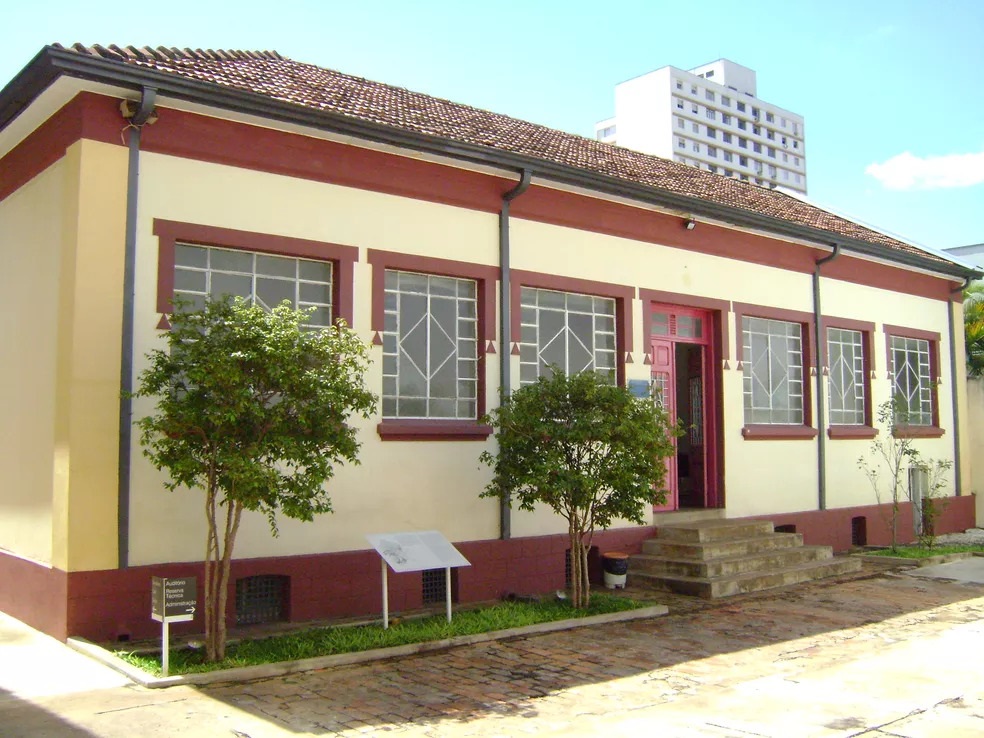 A imagem mostra um edifício histórico de um único andar, com arquitetura simples e tradicional. As paredes são de cor creme clara, com detalhes em vermelho escuro ao redor das janelas, da porta e na base do prédio. O telhado é feito de telhas de barro, típicas de construções antigas. A fachada tem cinco janelas com grades brancas em padrão geométrico, e uma porta central com moldura vermelha. Na frente do edifício, há um pequeno jardim com duas árvores jovens, um caminho pavimentado e um pequeno suporte com uma placa informativa. À direita da porta, há degraus que levam para dentro do prédio. Ao fundo, é possível ver um prédio moderno e alto, que contrasta com a simplicidade e o estilo antigo da construção em primeiro plano. O céu está azul e claro, indicando um dia ensolarado. Na fachada do edifício, há uma inscrição com as palavras “Auditório Reserva Técnica Administração”, sugerindo que o local é destinado a atividades administrativas e possivelmente culturais ou educativas. A construção transmite uma sensação de preservação histórica em meio a um ambiente urbano mais contemporâneo.