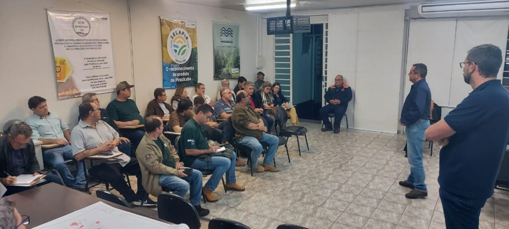 A imagem retrata uma reunião ou palestra realizada em uma sala fechada, onde um grupo de aproximadamente 15 pessoas está presente. * No lado direito da sala, um homem de camisa azul, de costas para a câmera, parece estar conduzindo a apresentação ou falando com o grupo. * À frente, sentado em uma cadeira isolada, há um homem vestindo um uniforme laranja e azul, possivelmente representando algum órgão de segurança ou defesa civil. * O restante dos participantes está sentado em cadeiras dispostas em fileiras, muitos deles fazendo anotações em cadernos ou observando atentamente. O público é majoritariamente masculino e veste trajes casuais. * Nas paredes do fundo, há cartazes informativos. Um deles menciona o "SIM" (Serviço de Inspeção Municipal) de Piracicaba, que certifica produtos de origem animal, e outro exibe o selo "SELAPIR", referente ao selo de inspeção local.