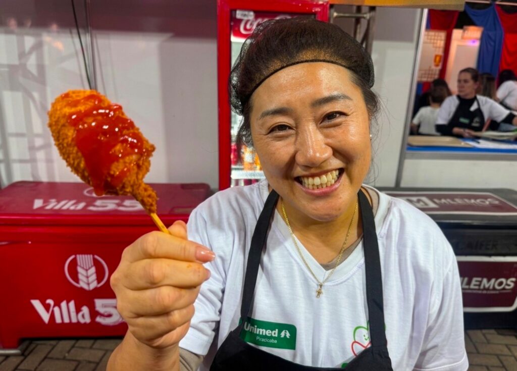 A imagem mostra uma mulher sorridente, vestindo uma camiseta branca com o logo "Unimed Piracicaba", um avental preto e uma touca para cabelo. Ela está segurando um salgado empanado em um palito, provavelmente um cachorro-quente ou algo similar, coberto com ketchup, e o apresenta em direção à câmera. Ao fundo, vê-se um ambiente que parece ser uma feira ou barraca de comida, com refrigeradores vermelhos e um freezer com a marca "Villa 55" e "MLEMOS". A mulher está em destaque, com uma expressão alegre e acolhedora, transmitindo simpatia e convidando o observador a experimentar o alimento. O cenário é iluminado e tem um ambiente informal e acolhedor.