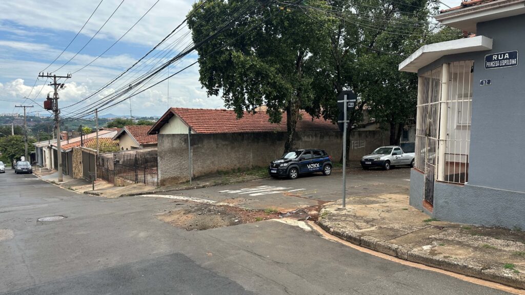 A imagem mostra uma rua residencial tranquila, com casas de um andar e telhados de cerâmica na cor terracota. A rua é pavimentada e apresenta algumas marcas de desgaste, além de uma tampa de bueiro visível no asfalto. No canto direito da imagem, há um prédio cinza com uma placa de rua que indica "RUA PRINCESA LEOPOLDINA" e o número 92. Na calçada ao lado do prédio, há um carro escuro estacionado com a inscrição "VOXX Vigilância" na lateral, indicando que provavelmente é um veículo de uma empresa de segurança. Ao lado do carro, está estacionada uma caminhonete branca. Ao fundo, uma parede clara delimita o terreno das casas, e uma árvore grande e frondosa se destaca, trazendo um pouco de verde ao cenário urbano. Fios de energia elétrica e postes cruzam a imagem, reforçando o ambiente típico de um bairro residencial. O céu está parcialmente nublado, com luz natural iluminando a cena. No geral, a imagem transmite a sensação de um bairro calmo e organizado.