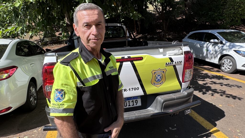 A imagem mostra um agente de trânsito em pé, posicionado à frente de uma caminhonete branca oficial. O agente veste um uniforme de alta visibilidade, predominantemente amarelo neon com detalhes pretos, incluindo faixas refletivas para maior segurança. A caminhonete atrás dele tem inscrições que indicam ser veículo oficial do departamento de trânsito da Prefeitura do Município de Piracicaba, com o número de identificação TR-1700 e a placa 0119. Ao redor, há outros carros estacionados e árvores ao fundo, sugerindo que a cena ocorre em um ambiente externo, possivelmente uma rua ou estacionamento. O agente está olhando diretamente para a câmera, transmitindo uma postura profissional e atenta. O ambiente está iluminado pela luz natural do dia, com sombras de árvores visíveis no chão.