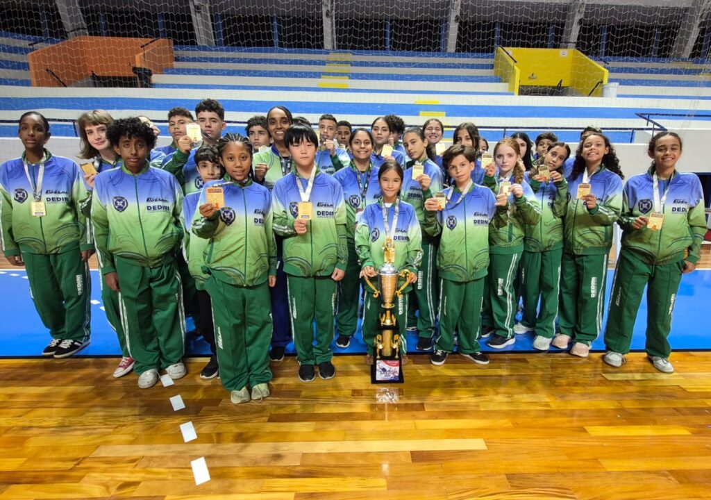 O arquivo mostra uma equipe infantojuvenil posando para comemorar uma vitória ou colocação num campeonato, possivelmente dentro de uma quadra poliesportiva. Detalhes visíveis: Participantes: muitas crianças e adolescentes (meninos e meninas) reunidos em grupo. Todos vestem uniformes combinando: jaquetas em degradê azul‑verde e calças verdes com o logotipo “DEDINI”. Troféu e medalhas: quase todos seguram medalhas douradas; uma criança no centro segura um troféu grande — sinal claro de que celebram um título ou um resultado importante. Ambiente: quadra com piso de madeira polida (com marcações no chão). Ao fundo aparecem arquibancadas nas cores azul e amarelo e redes de proteção, indicando um ginásio esportivo escolar ou comunitário. Atmosfera: bastante festiva e orgulhosa — expressão de conquista coletiva e espírito de equipe.