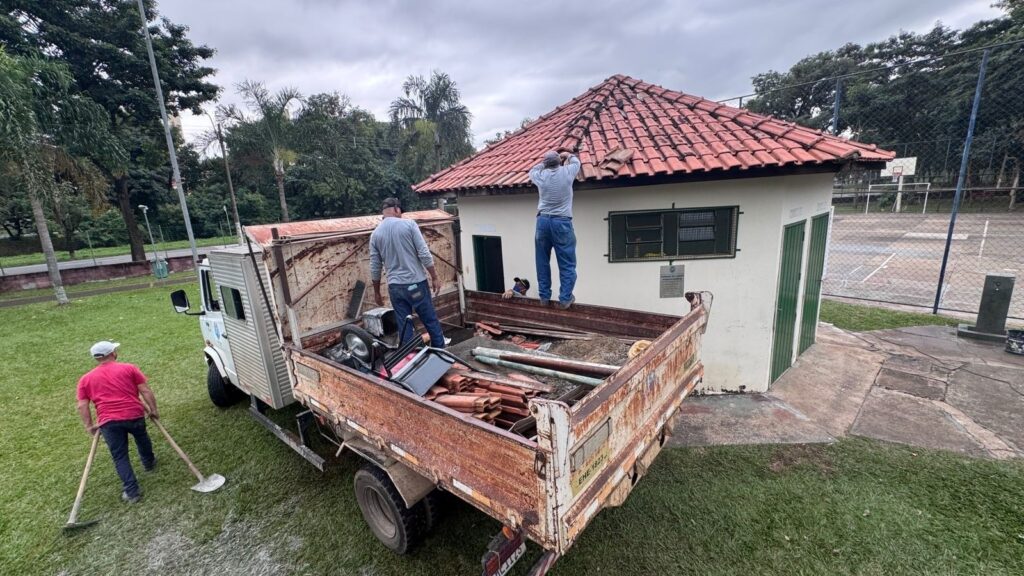 A imagem mostra três homens trabalhando na reforma ou manutenção de um pequeno prédio com telhado de telhas vermelhas. O prédio é claro, possivelmente branco ou bege, com uma janela protegida por grade metálica e uma porta verde. Um dos homens, vestido com uma camiseta rosa e boné branco, está no chão segurando uma ferramenta, possivelmente uma enxada ou enxadão. Os outros dois homens, ambos usando camisas cinza e calças jeans, estão sobre a caçamba de uma caminhonete branca, que parece estar carregando materiais de construção ou entulho. Um deles está organizando ou pegando materiais dentro da caçamba, enquanto o outro está trabalhando na beirada do telhado, possivelmente fazendo reparos. Ao fundo, é possível ver uma quadra de esportes cercada por uma cerca de arame. O céu está nublado, indicando um dia com pouca luz solar direta. A cena transmite uma atividade de manutenção em um espaço público.
