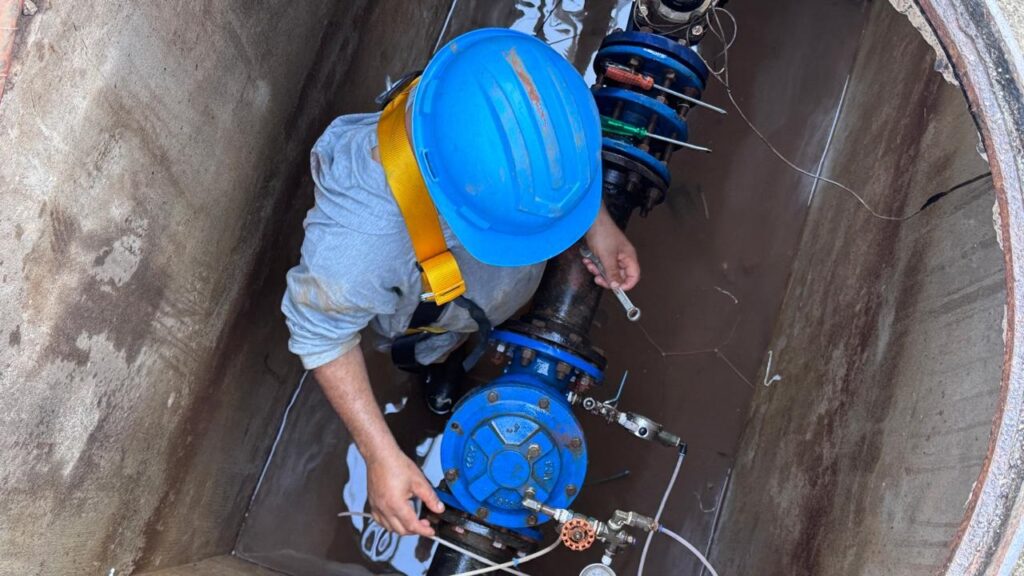 A imagem mostra uma visão aérea de um trabalhador dentro de uma estrutura subterrânea de concreto, possivelmente um poço de visita ou câmara de inspeção. * O profissional está equipado com um capacete de proteção azul e um cinto de segurança (arnês) amarelo, indicando o uso de EPIs para trabalho em espaço confinado. * Ele está realizando a manutenção ou reparo em uma tubulação hidráulica industrial, que possui flanges azuis e conexões metálicas. Com uma das mãos, ele manuseia uma chave de boca para ajustar parafusos ou componentes do sistema. * O ambiente apresenta um aspecto úmido com acúmulo de água no fundo da câmara, e é possível notar ferramentas, como chaves de fenda, deixadas sobre as flanges da tubulação.