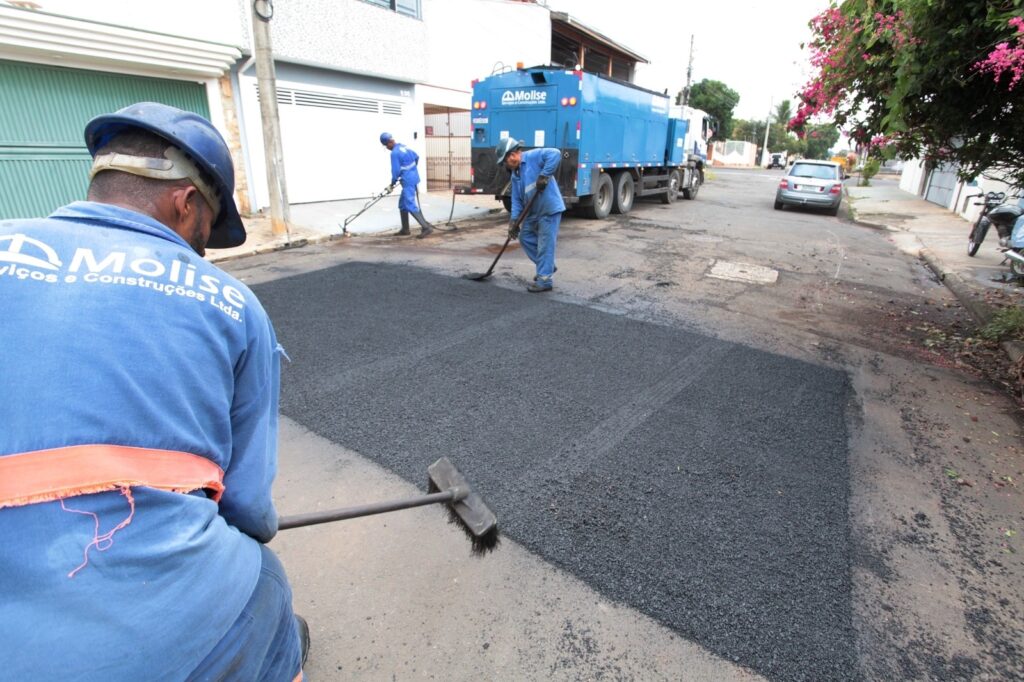 A imagem mostra uma equipe de três trabalhadores uniformizados da empresa "Molise Serviços e Construções Ltda." realizando o reparo ou a pavimentação de uma via pública. * O trabalhador em primeiro plano, visto de costas, utiliza uma vassoura para espalhar e nivelar o asfalto recém-aplicado. * Ao fundo, outros dois operários auxiliam no processo: um utiliza uma ferramenta manual para espalhar o material próximo à calçada, enquanto o outro utiliza uma espátula ou vassoura para dar o acabamento na massa asfáltica. * Um caminhão azul, também identificado com a marca da empresa, encontra-se estacionado na via, servindo de suporte para a operação. O cenário é uma rua residencial pavimentada, com casas nas laterais, uma árvore com flores rosas à direita e um veículo prateado estacionado ao fundo.