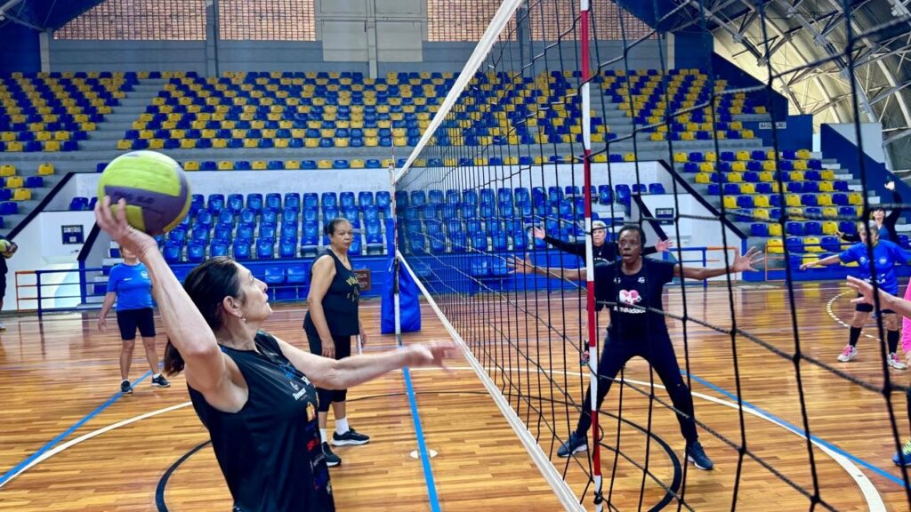 Esta imagem mostra um grupo de mulheres jogando vôlei em uma quadra coberta. * Em primeiro plano, à esquerda, uma jogadora prepara-se para sacar a bola, que está acima de sua cabeça. * Do outro lado da rede, uma jogadora com camisa preta está posicionada com os braços abertos, pronta para a defesa, enquanto outras jogadoras se espalham pela quadra. * O ambiente é uma quadra esportiva profissional com piso de madeira, rede de vôlei centralizada e arquibancadas com assentos nas cores azul e amarelo ao fundo. * A iluminação é artificial e o espaço parece bem conservado, sugerindo uma prática esportiva recreativa ou de treinamento.