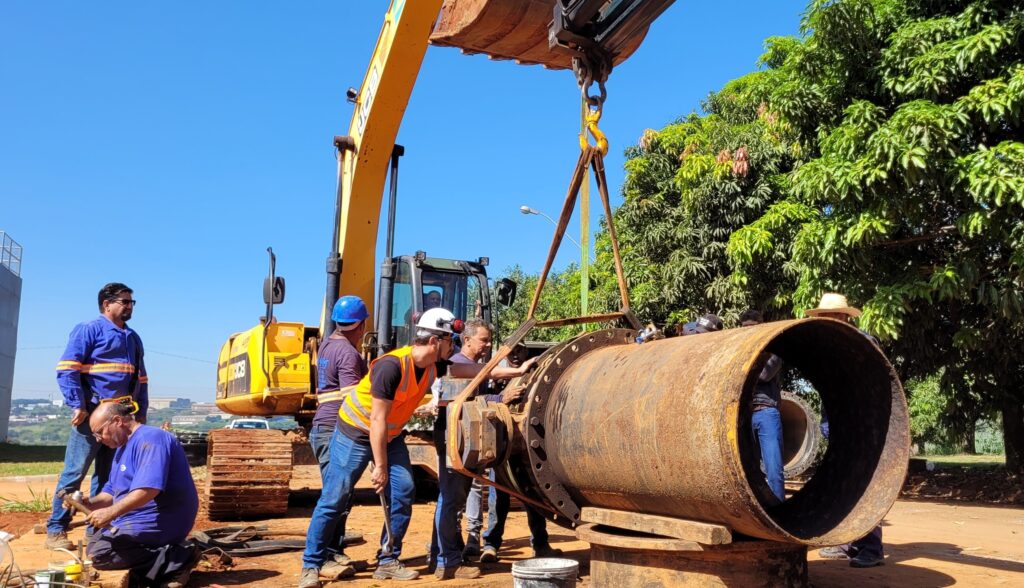 A imagem mostra uma cena industrial ao ar livre em um dia ensolarado. No centro, há uma escavadeira amarela da marca JCB, que está levantando e posicionando um grande tubo metálico enferrujado, preso por correntes ao acessório da máquina. Ao redor da escavadeira, várias pessoas estão trabalhando: um homem de camiseta azul está agachado no chão manipulando alguma ferramenta, enquanto outro, usando capacete branco e colete laranja de alta visibilidade, está ajudando a guiar o tubo. Ao fundo, mais trabalhadores também participam da tarefa. O cenário é um terreno de terra, com árvores verdes e céu azul ao fundo, indicando que o trabalho ocorre em uma área externa, possivelmente um canteiro de obras. A luz do sol é forte, criando sombras definidas no chão. A imagem destaca a colaboração entre os trabalhadores e o uso da máquina pesada para movimentar o material.