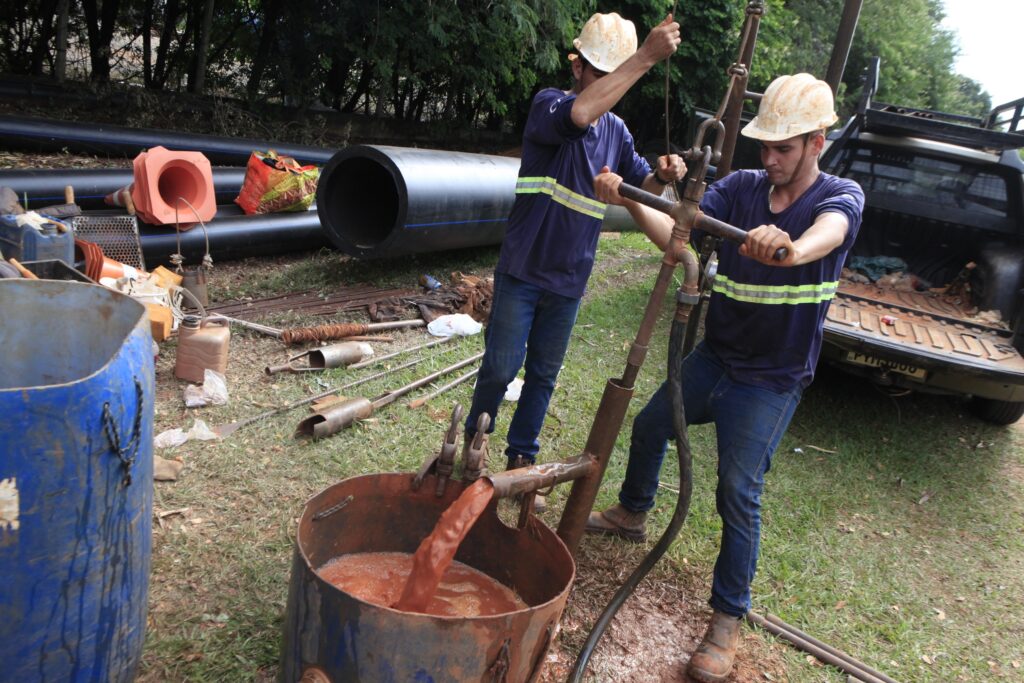 A imagem mostra dois homens trabalhando ao ar livre em um ambiente que parece ser uma obra ou um local de escavação. Ambos estão usando capacetes de segurança e camisas de trabalho com faixas refletivas, além de calças jeans. Um dos homens está puxando uma corda, enquanto o outro está operando uma máquina manual com alças. À frente deles, há um tambor metálico enferrujado, no qual está sendo bombeado um líquido de cor avermelhada, provavelmente por meio de um tubo conectado ao tambor. No fundo, é possível ver tubos grandes pretos espalhados pelo chão, cones de sinalização laranja e alguns materiais de construção. Também há uma caminhonete estacionada próxima. O cenário sugere um trabalho manual pesado, possivelmente relacionado a encanamento, drenagem ou algum tipo de manutenção industrial. O ambiente está iluminado pela luz natural do dia, com sombras visíveis. O chão é coberto por grama e terra, com algumas áreas aparentemente úmidas ou lamacentas. As cores predominantes são tons terrosos, azul dos tubos e do tambor, e o amarelo e verde das faixas refletivas nas roupas dos trabalhadores. A composição da imagem destaca os homens e o tambor como foco principal, enquanto o fundo contextualiza a atividade que está sendo realizada.