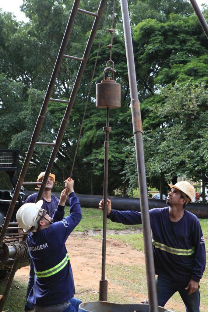 A imagem mostra três homens trabalhando ao ar livre com equipamentos de perfuração ou sondagem geotécnica. Dois dos homens estão em primeiro plano, ambos usando capacetes de segurança — um branco e outro bege — e uniformes azuis com faixas refletivas verdes e amarelas. O homem à esquerda, com capacete branco, está puxando uma corda amarrada a um peso metálico pesado, que está suspenso em uma estrutura metálica em forma de "A", enferrujada. O segundo homem, à direita e um pouco atrás, observa o peso suspenso, também segurando a corda. Um terceiro homem, parcialmente visível ao fundo, também usa capacete bege e observa a cena. O cenário é ao ar livre, com muitas árvores e vegetação ao fundo, sugerindo que o trabalho está sendo feito em uma área natural ou parque. O chão é de terra e grama. A iluminação indica que é durante o dia, com luz natural, possivelmente com sol filtrado pelas árvores. Na camisa do homem de capacete branco está escrito "ONDAGEM", indicando que o trabalho pode estar relacionado à sondagem do solo, que é um procedimento comum em engenharia para análise do terreno. A imagem transmite a ideia de trabalho em equipe e esforço físico na execução de uma tarefa técnica.