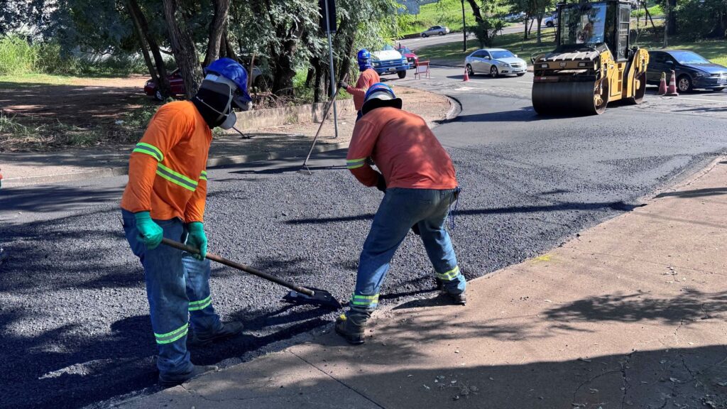 A imagem mostra uma cena de obras em uma estrada durante o dia. Dois trabalhadores estão no centro da imagem, ambos usando capacetes azuis e roupas de segurança laranja com faixas refletivas. Eles estão aplicando e nivelando asfalto fresco na via. Um dos trabalhadores está usando luvas verdes e ambos estão focados na tarefa manual de espalhar o asfalto. Ao fundo, há um terceiro trabalhador também com roupa de segurança laranja e capacete azul, segurando uma ferramenta longa. À direita, há uma máquina compactadora amarela da marca CAT, estacionada sobre o asfalto recém aplicado. Mais ao fundo, é possível ver uma rua com carros passando, calçadas, árvores e cones de sinalização laranja, indicando que a área está em obras. A iluminação é forte, sugerindo que a foto foi tirada em um dia ensolarado. A imagem representa um típico trabalho de manutenção ou pavimentação de estrada.