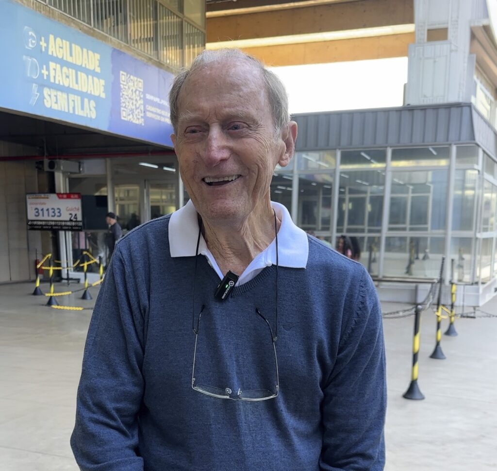 A imagem mostra um senhor de idade avançada, com cabelos grisalhos e um sorriso gentil, posicionado em primeiro plano. Ele veste um suéter azul com gola polo branca e utiliza um par de óculos pendurado por uma corrente ou cordão em seu pescoço, além de um pequeno microfone de lapela preso ao colarinho. Ao fundo, observa-se o ambiente de um estabelecimento público ou terminal de atendimento. Há uma placa azul fixada na parte superior com os dizeres "+ AGILIDADE + FACILIDADE SEM FILAS" e um código QR. Abaixo, vê-se um painel digital que exibe o número "31133" e indica "Guichê 24". O local parece ser organizado com delimitadores de fila amarelos e pretos, e há uma estrutura de vidro ao fundo, sugerindo um ambiente administrativo ou de serviços.