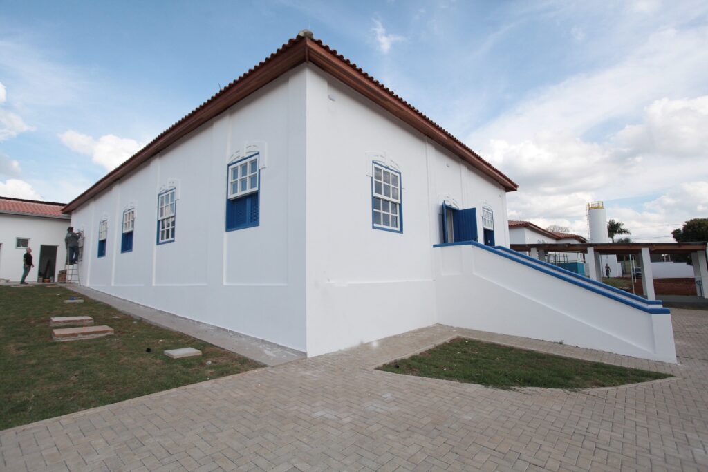 A imagem mostra um prédio colonial recém-reformado, pintado de branco com detalhes em azul nas janelas e portas. O telhado é de telhas cerâmicas vermelhas. O prédio tem dois andares, e há uma escada externa que leva ao segundo andar. No entorno, há uma área pavimentada e alguns trechos de grama. Dois trabalhadores aparecem na cena, um próximo a uma escada e outro mais distante, aparentemente realizando trabalhos de acabamento. Ao fundo, é possível ver um céu azul com algumas nuvens, além de uma torre de água e outra parte do conjunto de construções. A cena está bem iluminada pela luz natural do dia.
