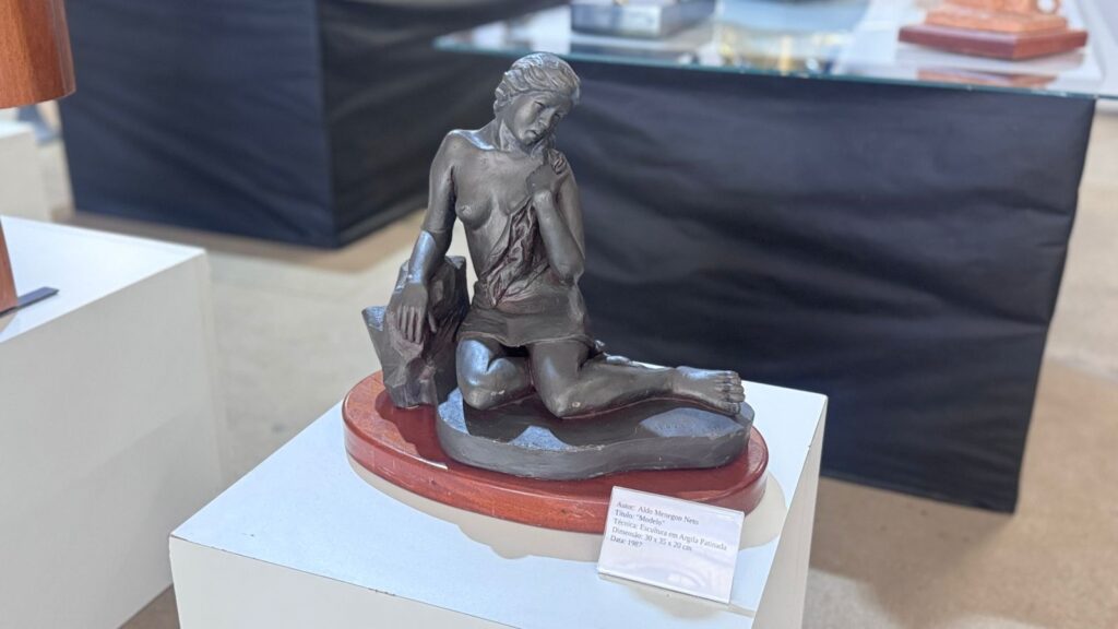 A imagem exibe uma escultura figurativa de bronze ou argila patinada, posicionada sobre uma base de madeira ovalada e um pedestal branco. A obra retrata uma figura feminina sentada em uma pose contemplativa, com uma das mãos apoiada em uma estrutura rochosa atrás de si e a outra segurando um tecido leve que cobre parte de seu corpo.
A estatueta apresenta detalhes realistas na anatomia e nas dobras da vestimenta. À frente da peça, há uma pequena placa informativa que detalha os dados da obra:
* Autor: Aldo Menegon Neto
* Título: "Modelo"
* Técnica: Escultura em Argila Patinada
* Dimensão: 30 x 35 x 20 cm
* Data: 1987
O cenário sugere uma exposição de arte ou galeria, com elementos desfocados ao fundo, como outras estruturas de exibição e superfícies de apoio, mantendo o foco central na escultura.