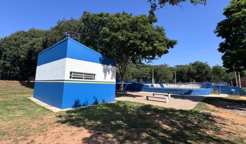 A imagem mostra uma cena ao ar livre em um dia ensolarado com céu azul claro. No centro da imagem há um pequeno prédio retangular pintado com listras horizontais em azul e branco, separado por uma faixa fina amarela e verde. O prédio possui uma janela horizontal com várias divisórias. À direita do prédio, há uma área que parece ser um skatepark, com rampas e paredes pintadas também em azul. Na frente do skatepark, há vários bancos simples e claros posicionados sobre um piso pavimentado. O chão ao redor é uma mistura de grama seca, terra e pavimento. Árvores verdes e densas cercam a área, oferecendo sombra em algumas partes da imagem. A perspectiva da foto é ligeiramente baixa, olhando para cima em direção ao prédio e ao skatepark, com as árvores ao fundo contrastando com o céu. A composição transmite uma sensação de espaço moderno e organizado, dedicado ao lazer ao ar livre.