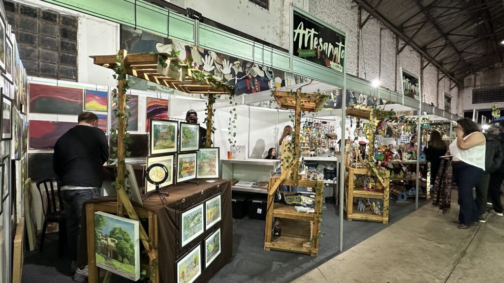 A imagem mostra um mercado de artesanato em um ambiente interno, provavelmente um galpão com paredes de tijolos pintados de branco e teto alto com estrutura metálica. Há várias barracas organizadas em fileira, todas com uma placa verde no topo que diz "Artesanato Piracicabano". As barracas são feitas com estrutura de madeira clara e decoradas com folhagens artificiais, dando um aspecto natural e acolhedor. Na barraca em primeiro plano, há várias pinturas emolduradas apoiadas sobre uma mesa coberta com um tecido marrom. Também é possível ver uma luminária circular de luz LED, provavelmente usada para iluminar melhor os produtos expostos. Um homem com boné preto está atrás da mesa, possivelmente o artesão ou vendedor. Ao redor, algumas pessoas observam os produtos: um homem com camiseta preta está à esquerda olhando as pinturas, uma mulher com blusa branca e calça jeans está à direita, também observando os itens. No fundo, outras barracas exibem objetos variados, como pequenas esculturas ou peças decorativas. O ambiente transmite uma atmosfera tranquila e convidativa, típica de feiras de artesanato onde artistas locais exibem e vendem suas criações.