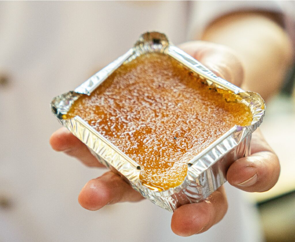 A imagem mostra uma mão humana segurando uma pequena forma retangular de alumínio descartável. Dentro da forma, há um doce de cor alaranjada ou dourada, com uma textura granulada ou cristalizada na superfície, lembrando um doce de leite, uma cocada assada ou um pudim de consistência firme. A iluminação é suave e destaca o brilho da superfície do doce e o reflexo metálico da forma de alumínio. O fundo está desfocado, sugerindo um ambiente de cozinha ou confeitaria.