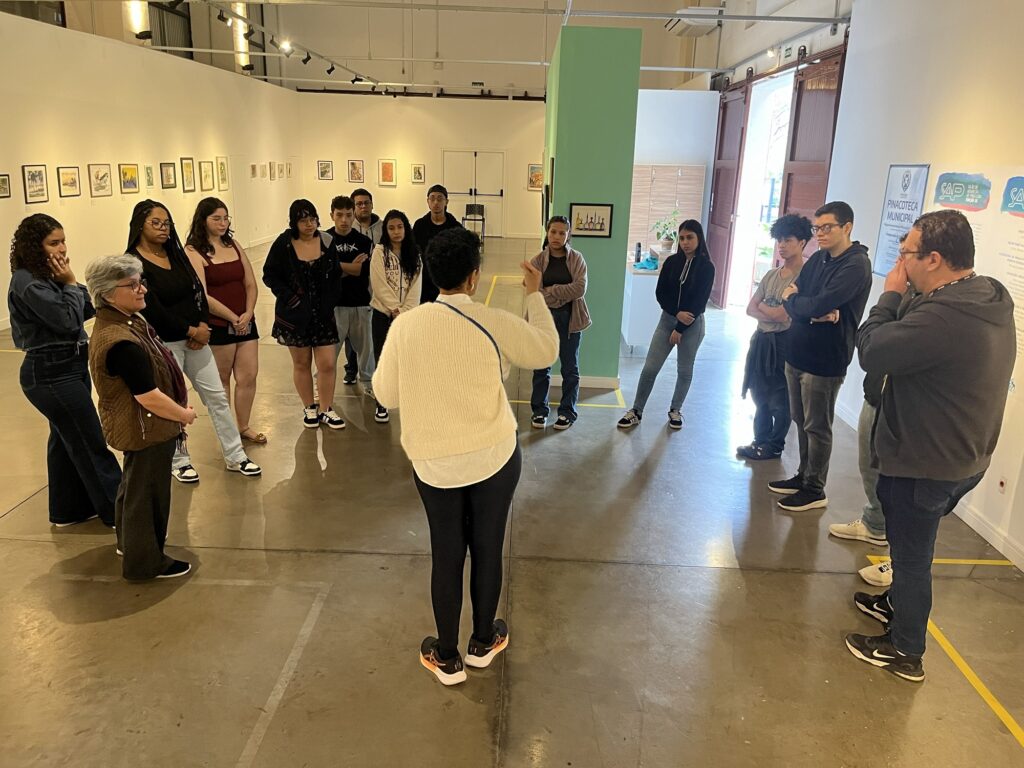 A imagem mostra um grupo de jovens em uma visita guiada a uma galeria de arte ou pinacoteca. * No centro, de costas, uma mulher com um suéter bege gesticula enquanto explica algo para o grupo, que a observa atentamente. * O grupo é composto por cerca de 13 pessoas de diferentes idades e estilos, dispostas em um semicírculo ao redor da guia. * O espaço é uma galeria moderna e bem iluminada, com paredes brancas decoradas com diversas obras de arte emolduradas. * No chão, há fitas adesivas amarelas demarcando o espaço, e ao fundo, destaca-se uma parede pintada de verde-água. À direita, é possível ver um cartaz informativo sobre o "Salão de Artes de Piracicaba".