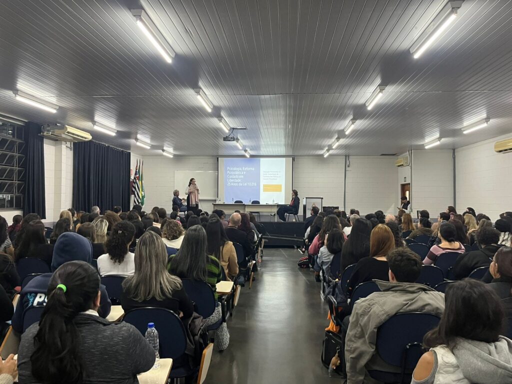 A imagem mostra um auditório amplo e bem iluminado, ocupado por uma plateia numerosa de pessoas sentadas em cadeiras azuis, voltadas para o palco. No palco, há uma mesa com dois palestrantes e um projetor exibindo um slide com o título: "Psicologia, Reforma Psiquiátrica e Cuidado em Liberdade: 25 Anos da Lei 10.216". O ambiente possui teto com várias luminárias lineares, paredes brancas e cortinas escuras ao fundo, criando uma atmosfera de evento acadêmico ou profissional. A perspectiva da foto é tirada do fundo da sala, oferecendo uma visão completa da audiência e da apresentação.