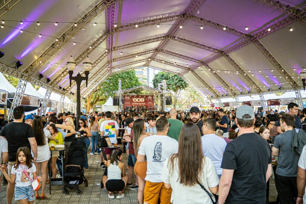 A imagem retrata um festival de comida de rua ao ar livre, realizado sob uma grande estrutura de tenda metálica decorada com luzes suspensas. O ambiente está repleto de pessoas de diversas idades, criando uma atmosfera social e vibrante. * No centro da imagem, há uma multidão concentrada, com muitas pessoas em pé, conversando, bebendo e aproveitando o evento. * Ao fundo, é possível ver um palco onde ocorre uma apresentação musical, com o nome do evento "Comida na Rua" em destaque em um painel. * Em primeiro plano, vemos pessoas de costas e de lado, incluindo uma criança à esquerda usando uma camiseta branca, e um carrinho de bebê, sugerindo um ambiente familiar. * A iluminação é composta por tons quentes das luzes de corda e focos de luz roxa na estrutura do teto, contrastando com a luz natural do final da tarde que entra pelas laterais da tenda.