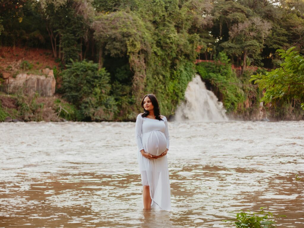 A imagem mostra uma mulher grávida em pé dentro de um rio, com água até a metade das pernas. Ela veste um vestido branco longo e fluido, que destaca a barriga da gravidez, a qual ela segura com as duas mãos. A mulher está de perfil, olhando para a direita, com uma expressão serena e contemplativa. Ao fundo, há uma cachoeira caindo sobre pedras, cercada por uma vegetação verde e densa, sugerindo um ambiente natural e tranquilo. A luz natural ilumina suavemente a cena, criando um contraste harmonioso entre o branco do vestido, o marrom da água do rio e o verde das plantas ao redor. A composição da imagem valoriza a conexão entre a mulher e a natureza ao seu redor.