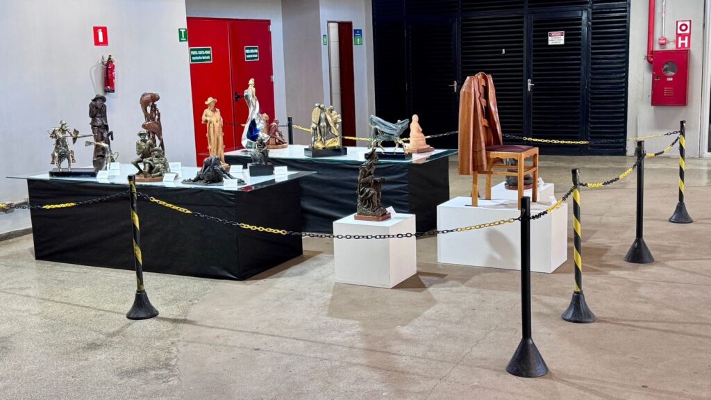 A imagem apresenta uma exposição de arte contemporânea instalada em um ambiente interno, possivelmente um saguão ou galeria. * No centro, há duas mesas cobertas com tecidos pretos que sustentam diversas esculturas de diferentes tamanhos, materiais e estilos, variando entre figuras humanas, formas abstratas e representações de animais ou objetos. * À direita, destaca-se uma instalação composta por uma cadeira de madeira sobre um pedestal branco, com um casaco de couro marrom pendurado no encosto, sugerindo uma representação da ausência humana. * O espaço é delimitado por uma corrente de segurança amarela e preta sustentada por pedestais metálicos, criando uma barreira entre o público e as obras. * O ambiente possui um estilo industrial, com piso de concreto, portas corta-fogo vermelhas ao fundo, sinalização de segurança (extintores e placas de saída) e portas metálicas pretas, indicando que a exposição está montada em um espaço funcional.