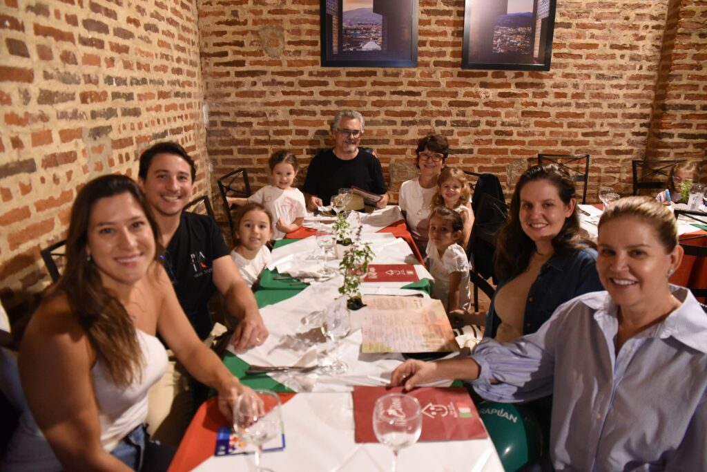 A imagem mostra um grupo de dez pessoas reunidas ao redor de uma mesa longa em um restaurante. Parece ser uma reunião familiar, com adultos e várias crianças de diferentes idades. A mesa está decorada com toalhas brancas e caminhos de mesa em vermelho e verde, além de pequenos vasos com plantas como centro de mesa. Todos estão sentados, sorrindo e parecem felizes, criando uma atmosfera acolhedora e alegre. O ambiente do restaurante tem uma decoração rústica, com paredes de tijolos aparentes e duas fotos emolduradas penduradas na parede ao fundo, mostrando paisagens naturais. A iluminação é quente, contribuindo para o clima intimista do encontro. Um balão verde com a palavra "RAPLAN" está visível próximo à mesa, sugerindo que pode ser uma comemoração ou evento especial. No geral, a imagem transmite um momento de confraternização e alegria entre familiares ou amigos em um ambiente confortável e agradável.