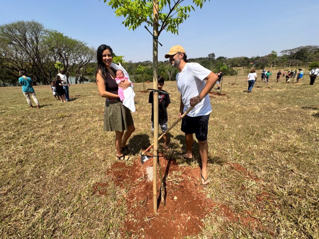 A imagem mostra uma cena ao ar livre durante um evento de plantio de árvores em um dia ensolarado. No centro da imagem, há uma família composta por quatro pessoas. À esquerda, uma mulher de cabelos longos e escuros está segurando um bebê pequeno enrolado em um cobertor branco. Ao lado dela, um menino está em pé, usando uma camiseta preta, próximo a uma muda recém-plantada. Um homem, vestindo camiseta branca, bermuda escura, boné bege e chinelos, está usando uma pá para ajeitar a terra ao redor da muda, que está apoiada por uma estaca de bambu alta. O chão é coberto por uma grama seca e amarelada, com algumas áreas de terra exposta de cor avermelhada, onde as árvores foram plantadas. Ao fundo, é possível ver um campo aberto com algumas árvores espalhadas, um corpo d’água e outras pessoas participando do plantio, indicando que se trata de uma iniciativa comunitária voltada para o meio ambiente. A luz é natural e intensa, típica do meio-dia, criando sombras suaves. A foto é feita com uma lente grande-angular, mostrando bem o contexto da atividade.