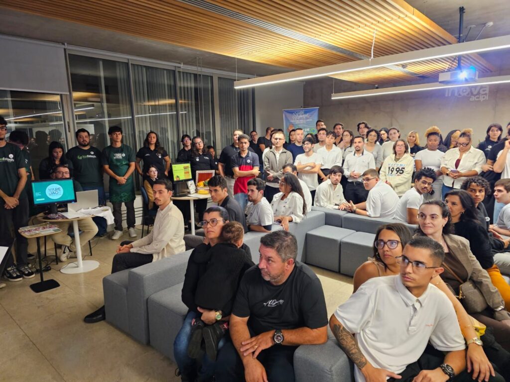 A imagem mostra um ambiente interno moderno, possivelmente uma sala de eventos ou um espaço de inovação, onde um grupo grande de pessoas está reunido. A maioria delas está sentada em sofás modulares cinza, enquanto outras estão em pé ao fundo, todas voltadas para a esquerda, em direção a uma apresentação ou palestra. Na parte esquerda da imagem, há uma mesa com um monitor que exibe a palavra "VAREJÃO" e um laptop. Próximo a essa mesa, algumas pessoas estão usando aventais verdes com a marca "Inofatech". O ambiente tem um teto alto com iluminação linear e detalhes em madeira, além de paredes de concreto. Na parede à direita, há um logo que diz "inova acipi". O público é composto principalmente por jovens adultos, vestidos de maneira casual e informal. Em primeiro plano, uma mulher segura uma criança pequena no colo. O clima geral parece ser profissional, porém descontraído, indicando que pode estar ocorrendo uma palestra, workshop ou evento comunitário voltado para inovação ou empreendedorismo.