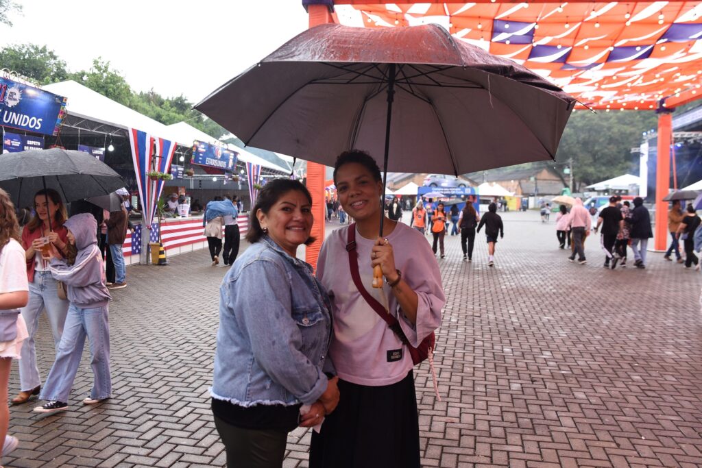 A imagem mostra duas mulheres sorrindo sob um guarda-chuva cinza escuro em um ambiente ao ar livre, aparentemente durante uma leve chuva. Elas estão próximas uma da outra, com a mulher à direita segurando o guarda-chuva. A mulher à esquerda tem cabelos escuros, veste uma jaqueta jeans e calça preta, enquanto a mulher à direita usa uma blusa clara, saia preta, e uma bolsa vermelha atravessada no corpo, além de estar usando aparelho nos dentes. Ao fundo, há barracas decoradas com as cores da bandeira dos Estados Unidos e placas com palavras em português, como "FESTA DAS NAÇÕES", "ESTADOS UNIDOS", "ESPETO DE FRANGO" e "LANCHE". O local parece ser uma praça pavimentada, com algumas pessoas circulando e outras segurando guarda-chuvas. Há também tecidos coloridos (laranja e azul) pendurados acima, possivelmente como parte da decoração do evento. A atmosfera sugere uma celebração cultural ou festival de nações, com clima nublado e chão molhado pela chuva.