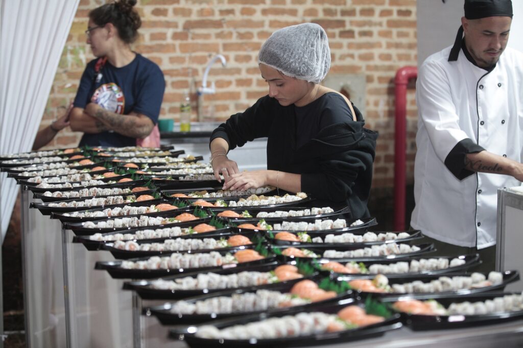 A imagem mostra uma cozinha profissional onde várias pessoas estão preparando sushi em grande quantidade. No centro, uma mulher jovem, vestindo uma blusa preta de mangas longas e uma touca branca, está organizando pedaços de sushi em uma bandeja preta longa e estreita. Ao lado dela, à direita, um chef de cozinha com uniforme branco e boné preto também está envolvido na preparação do sushi. À esquerda, uma mulher de óculos e tatuagens nos braços observa o processo com os braços cruzados. Na frente deles, há muitas bandejas pretas alinhadas, todas cheias de sushi, estendendo-se em direção ao fundo da cozinha. O ambiente tem uma parede de tijolos expostos, uma pia de aço inoxidável e um cano vermelho aparente no teto, com iluminação clara que destaca os detalhes dos alimentos e do local. A cena transmite uma sensação de trabalho em linha de montagem, com foco na organização e produção em massa dos sushis.