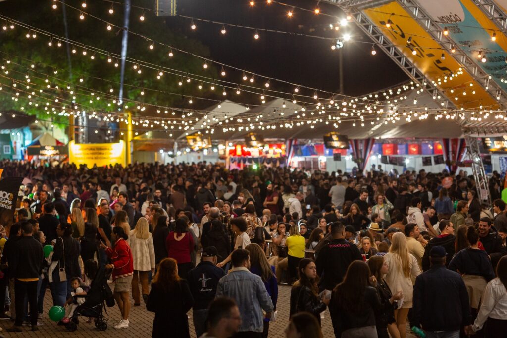 A imagem mostra uma cena noturna vibrante de um grande evento ao ar livre, possivelmente um festival ou feira, com uma enorme quantidade de pessoas reunidas. O ambiente está iluminado por várias fileiras de luzes penduradas no alto, que criam um efeito de teto luminoso e uma atmosfera acolhedora e festiva. No centro da imagem, há uma multidão densa, com pessoas de diferentes idades e estilos, algumas conversando, outras caminhando entre as barracas e tendas que aparecem ao fundo. Entre os elementos visíveis, destaca-se um carrinho de bebê e balões verdes, além de uma pessoa vestindo uma jaqueta preta com a palavra "Produção" nas costas, indicando que faz parte da equipe do evento. No lado esquerdo, há um banner com os dizeres "100 YEARS CATERPILLAR", sugerindo que a ocasião pode ser uma celebração do centenário da empresa Caterpillar, possivelmente patrocinadora do evento. A imagem transmite uma sensação de movimento, agitação e confraternização, com um clima alegre e descontraído sob a iluminação amarelada das lâmpadas. O chão é pavimentado, e a composição da foto é feita em ângulo amplo, capturando tanto o público quanto as estruturas temporárias montadas para a festa, como tendas e banners. O céu escuro contrasta com a iluminação intensa do evento, realçando o caráter noturno da celebração.