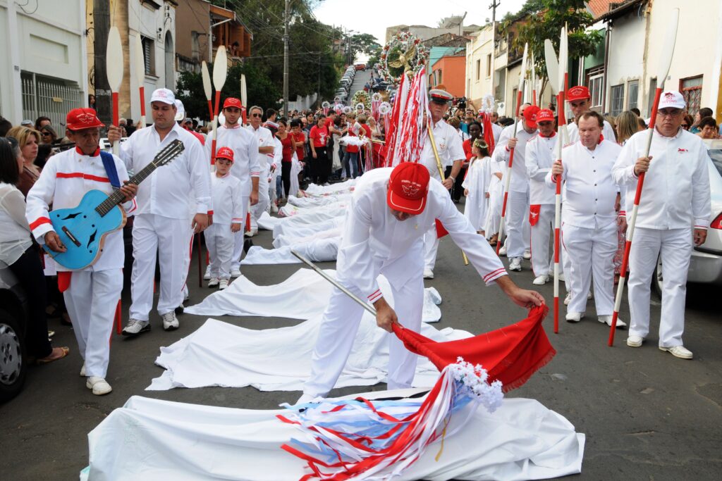 A imagem mostra uma procissão religiosa ao ar livre, provavelmente durante a "Festa do Divino", um evento tradicional em países de língua portuguesa. No centro da cena, vários homens vestem uniformes brancos com detalhes em vermelho, incluindo bonés vermelhos. Alguns carregam objetos longos e brancos com pontas vermelhas, que parecem remos ou estandartes. Um dos participantes toca um violão azul vibrante, enquanto outro segura uma bandeira vermelha com fitas brancas e azuis, posicionando-a sobre um lençol branco estendido no chão. Outros lençóis brancos também estão espalhados pela rua, indicando um ritual ou cerimônia específica do evento. Ao fundo, é possível ver prédios, carros estacionados e uma multidão de espectadores acompanhando a procissão. A atmosfera parece solene e festiva ao mesmo tempo, com a predominância das cores branca e vermelha, que são símbolos marcantes da celebração. A imagem transmite um forte senso de tradição, fé e comunidade.