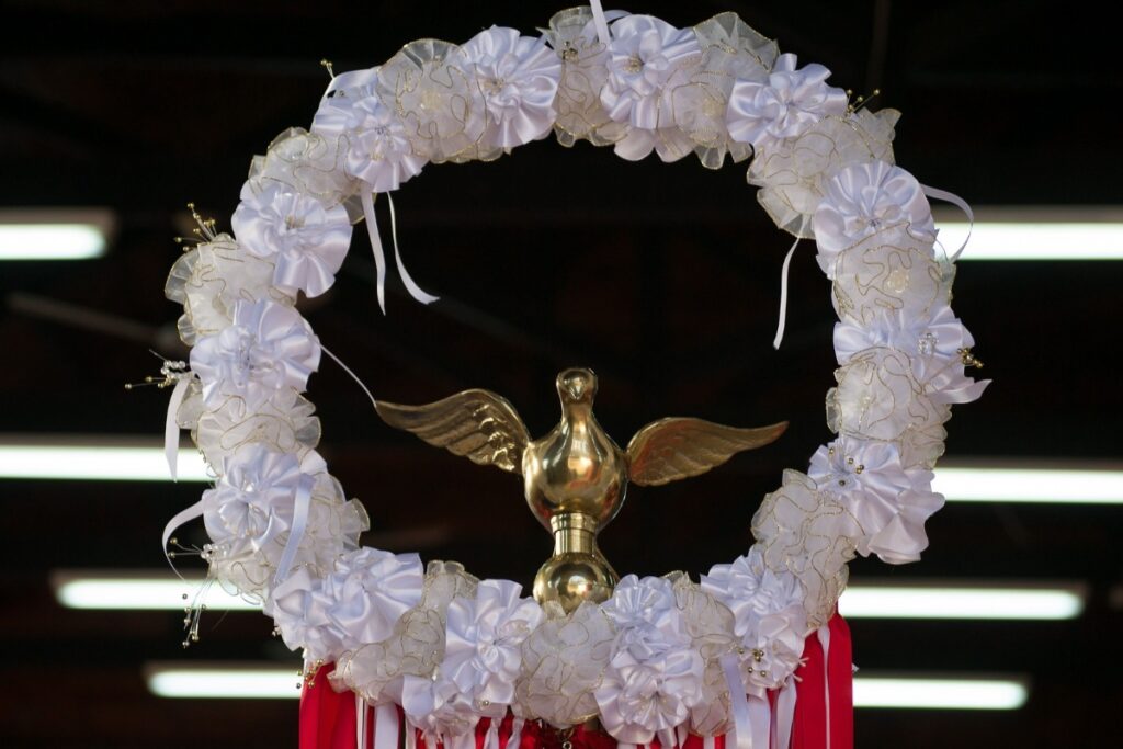 A imagem mostra uma peça decorativa central composta por uma coroa circular feita de flores brancas de tecido e fitas translúcidas com bordas douradas. No centro da coroa, há uma escultura dourada de um pombo em voo, com as asas abertas, simbolizando paz ou o Espírito Santo. A escultura está apoiada em um pequeno pedestal. Abaixo da coroa, pendem fitas vermelhas e brancas. O fundo é escuro e desfocado, com algumas linhas horizontais de luz branca, possivelmente de lâmpadas fluorescentes, que contrastam com a peça iluminada. A imagem transmite uma sensação de solenidade e espiritualidade.