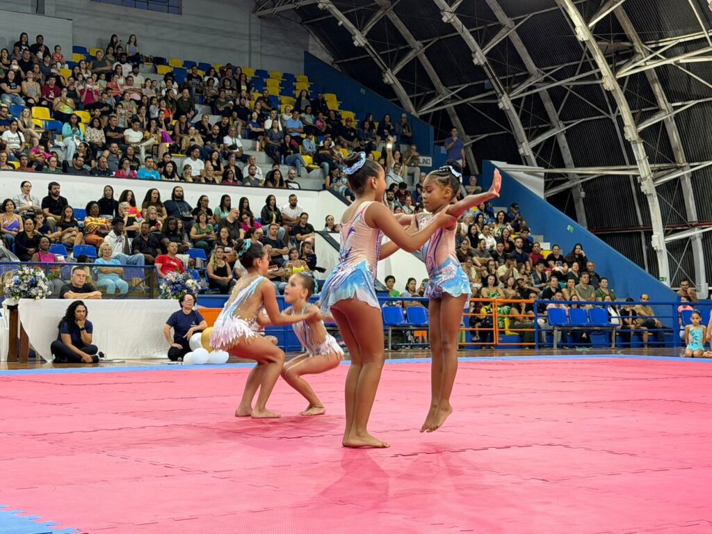 A imagem mostra uma apresentação de ginástica rítmica em um ginásio coberto, com um tapete rosa vibrante no centro. Quatro meninas jovens estão realizando uma coreografia, vestindo roupas de ginástica coloridas em tons de rosa, branco e azul, com detalhes delicados e saias rodadas. Duas delas estão em pé, uma segurando a outra no ar, enquanto as outras duas estão agachadas, formando uma composição harmoniosa. Seus cabelos estão presos em coques com acessórios combinando. Ao fundo, há uma arquibancada cheia de espectadores de várias idades, observando atentamente a apresentação. O teto é alto, com estrutura metálica aparente, e as paredes são azuis, características típicas de um ginásio esportivo. Também é possível ver uma mesa decorada com flores e uma pessoa sentada próxima a ela, possivelmente uma juíza ou organizadora do evento. A iluminação é clara, destacando bem as atletas e o ambiente ao redor.