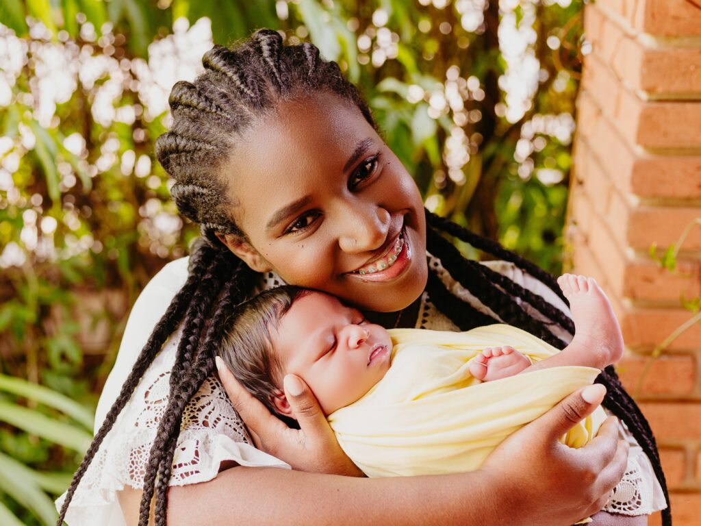 A imagem mostra uma mulher negra sorridente segurando um bebê recém-nascido enrolado em um cobertor amarelo. A mulher tem cabelos trançados e está usando aparelho nos dentes, com um sorriso aberto e olhar direto para a câmera. O bebê está dormindo tranquilamente, encostado no ombro da mulher. O fundo é desfocado, com elementos naturais como plantas e uma parede de tijolos, sugerindo que a foto foi tirada ao ar livre. A luz natural destaca os rostos e transmite uma sensação de carinho e ternura entre mãe e filho.