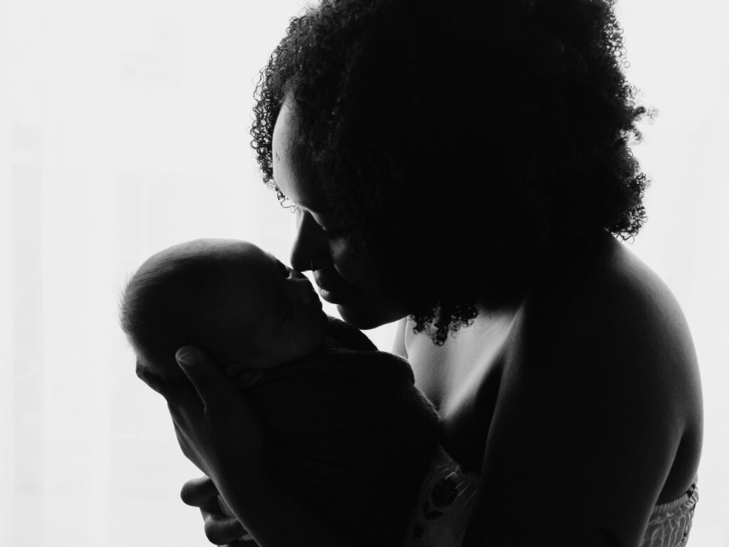 A imagem mostra uma silhueta em preto e branco de uma mãe segurando seu bebê recém-nascido próximo ao rosto. A mãe está inclinada para baixo, com o nariz quase tocando a testa do bebê, transmitindo um momento de carinho e conexão profunda entre eles. O fundo é claro e difuso, provavelmente uma janela, o que destaca as formas escuras da mãe e do bebê. A mãe tem cabelos cacheados visíveis na parte superior da imagem, e o bebê está enrolado em um cobertor, protegido nos braços da mãe. A composição transmite tranquilidade, amor e intimidade, capturando a essência do vínculo entre mãe e filho.