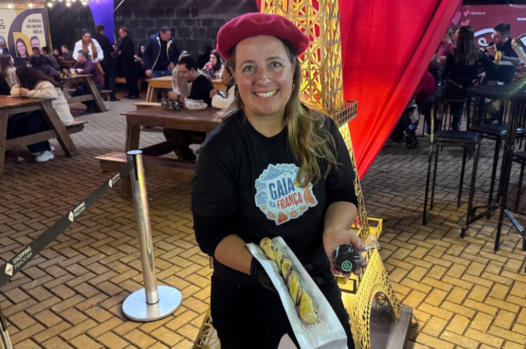 A imagem mostra uma mulher sorridente em um ambiente externo à noite, possivelmente em um evento temático sobre a França. Ela está vestindo um boina vermelha, uma blusa preta com o logo "GAIA NA FRANÇA" e luvas pretas. Na mão direita, ela segura um tipo de pão ou massa enrolada, enquanto na mão esquerda segura um pequeno doce embrulhado. Ao fundo, há uma estrutura iluminada que lembra a Torre Eiffel, reforçando o tema francês do evento. Também é possível ver mesas e cadeiras de madeira com pessoas sentadas, sugerindo um ambiente de feira ou festival gastronômico. À esquerda, há uma fita com a inscrição "SHOPPING PIRACICABA", indicando o local ou patrocinador do evento. A iluminação é quente e cria uma atmosfera acolhedora e festiva.