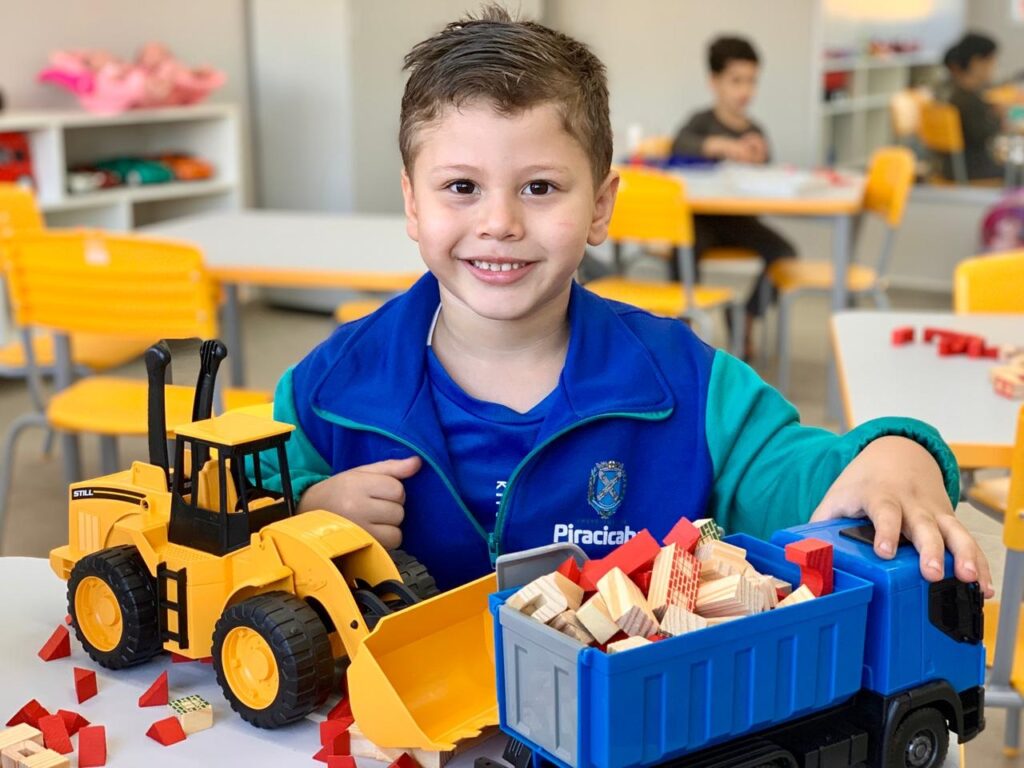 A imagem apresenta um menino sorridente em primeiro plano, sentado em uma sala de aula ou ambiente escolar. Ele veste uma jaqueta azul e verde, com um emblema escolar visível no peito. * O menino está brincando sobre uma mesa branca com dois grandes veículos de brinquedo: um trator amarelo (pá carregadeira) à esquerda e um caminhão basculante azul à direita. * A caçamba do caminhão está cheia de pequenos blocos de construção de madeira e peças vermelhas, que também estão espalhadas pela mesa ao redor dos brinquedos. * Ao fundo, em um ambiente desfocado, é possível ver outras mesas, cadeiras amarelas e outro aluno sentado, sugerindo um cenário típico de uma escola infantil ou creche. A iluminação é clara e o ambiente parece organizado e lúdico.
