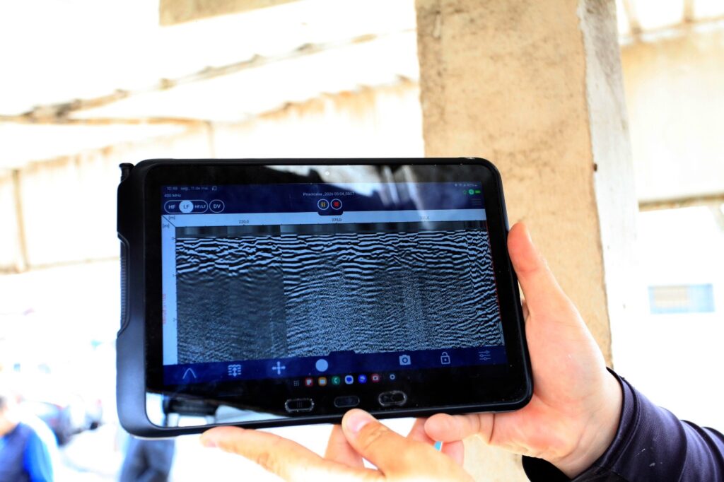 A imagem mostra uma pessoa segurando um tablet robusto, possivelmente à prova de impactos, com as duas mãos. Na tela do tablet, está exibida uma imagem de um radar de penetração no solo (GPR), que apresenta padrões em preto e branco, indicando camadas subterrâneas ou varreduras geofísicas. No topo da tela, há algumas informações técnicas, como a frequência de 400 MHz, opções de frequência (HF, LF, HF/LF, DV) e uma referência de arquivo "Piracicaba_2026 05 04_0007". A interface do software parece ser específica para análise geofísica ou de inspeção do solo. O ambiente ao fundo está desfocado, mas sugere um local externo ou industrial, com iluminação natural. A imagem transmite uma sensação de tecnologia aplicada em campo para estudos do subsolo.