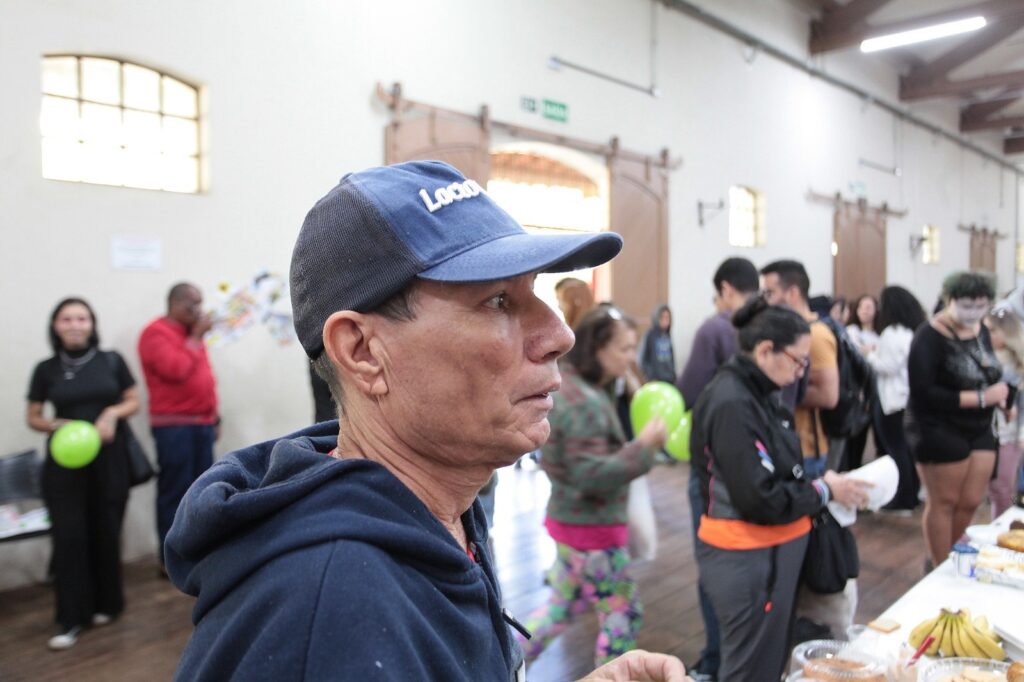 Esta imagem registra um momento de confraternização ou evento social em um ambiente interno amplo, com piso de madeira e paredes claras. * Em primeiro plano, um homem de perfil, vestindo um moletom azul escuro e um boné azul com a inscrição "Loca", olha para o lado direito. * O ambiente está movimentado com diversas pessoas circulando. À esquerda, uma mulher de preto segura um balão verde, enquanto um homem de camisa vermelha está ao fundo. * À direita, um grupo de pessoas interage perto de uma mesa onde há uma oferta de alimentos, incluindo bananas e outros itens, sugerindo um café da manhã ou lanche coletivo. * A iluminação é natural, vinda de janelas altas, e o ambiente transmite um clima informal e comunitário.