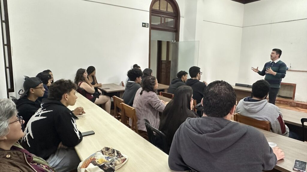 A imagem mostra uma sala de aula ou auditório com um grupo de alunos sentados em mesas longas, voltados para um instrutor que está em pé à direita, gesticulando enquanto fala. * O instrutor, um homem de meia-idade com barba, veste uma camisa listrada em tons de verde e azul. Ele ocupa uma posição de destaque, conduzindo a apresentação ou aula. * Os alunos estão dispostos em fileiras, com perfis diversos. Alguns estão atentos ao palestrante, enquanto outros olham para direções diferentes. Há uma mistura de jovens e adultos, vestindo roupas casuais como moletons e camisas. * O ambiente possui paredes brancas, piso de madeira e uma porta de arco ao fundo, sugerindo uma arquitetura clássica ou institucional. A iluminação é clara e uniforme, típica de um espaço de aprendizado. * Sobre a mesa em primeiro plano, à esquerda, nota-se uma bolsa estampada e um celular, indicando o uso cotidiano do espaço pelos estudantes.