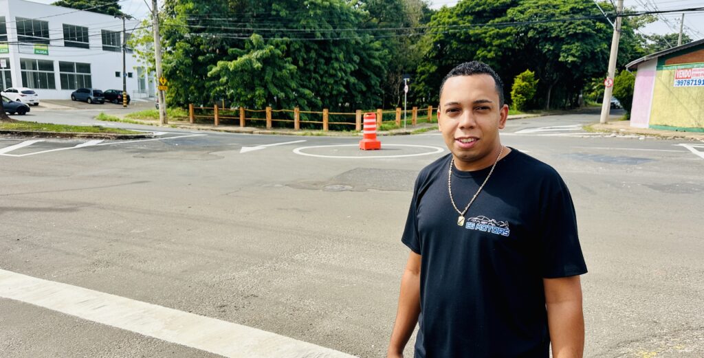 A imagem mostra um homem posando em uma rua, próximo a um cruzamento. Ele está no centro da imagem, vestindo uma camiseta preta com o logotipo "EG MOTORS" estampado na frente, além de usar um cordão dourado no pescoço. O homem tem cabelo curto e está sorrindo levemente para a câmera. Ao fundo, vê-se um cruzamento com uma área circular branca pintada no chão, onde há um cone de trânsito laranja e branco no centro. Há também uma cerca baixa de madeira e várias árvores verdes ao redor, sugerindo que a área é arborizada. Na parte esquerda da imagem, há um prédio branco com placas de "ALUGA", indicando que o local está disponível para aluguel. Do lado direito, há outra construção com uma placa de "VENDO" e um número de telefone. O céu está nublado, indicando um dia com pouca luz solar direta. No geral, a cena parece ser em uma área urbana tranquila, com o homem em destaque na frente do cenário.