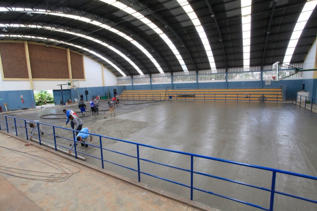 A imagem mostra um grupo de trabalhadores em uma quadra esportiva coberta, realizando a instalação de um piso de concreto. O ambiente é um ginásio com teto arqueado, feito de painéis translúcidos que permitem a entrada de luz natural, iluminando o local de forma suave. As paredes são azuis e há uma seção de tijolos aparentes à esquerda. Do outro lado da quadra, há arquibancadas amarelas em degraus. No centro da imagem, o piso de concreto está sendo nivelado e alisado por vários trabalhadores que usam ferramentas longas, como régua de alumínio para espalhar o concreto. Eles vestem roupas de trabalho simples, e alguns usam chapéus. A foto foi tirada de uma posição elevada, atrás de uma grade azul, o que oferece uma visão ampla da quadra e do processo de construção em andamento. A cena transmite uma sensação de trabalho manual intenso e cuidado na execução da obra, com destaque para a luminosidade natural que realça a superfície fresca do concreto.