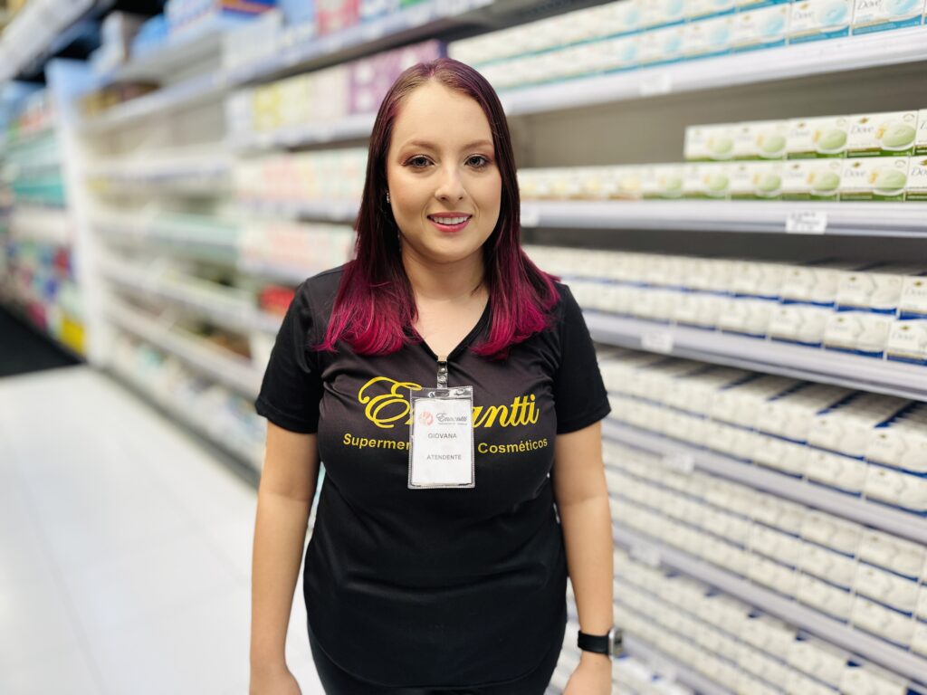 A imagem mostra uma jovem mulher em um supermercado, mais especificamente na seção de cosméticos. Ela está sorrindo e olhando diretamente para a câmera, transmitindo simpatia e profissionalismo. A mulher veste uma camiseta preta com o logo e o nome "Enanti Supermercado de Varejo" em amarelo, além de um crachá transparente preso à camiseta, indicando que ela é funcionária do local. Seu cabelo é escuro com mechas em tom roxo vibrante, e ela está usando maquiagem, incluindo sombra nos olhos e batom. Ao fundo, há prateleiras cheias de produtos, principalmente sabonetes em embalagens brancas e azuis, embora estejam desfocados para destacar a pessoa em primeiro plano. A iluminação é clara e uniforme, típica de ambientes internos comerciais. A composição da foto foca na atendente, centralizando-a na imagem, com as prateleiras criando um ambiente que contextualiza seu local de trabalho.
