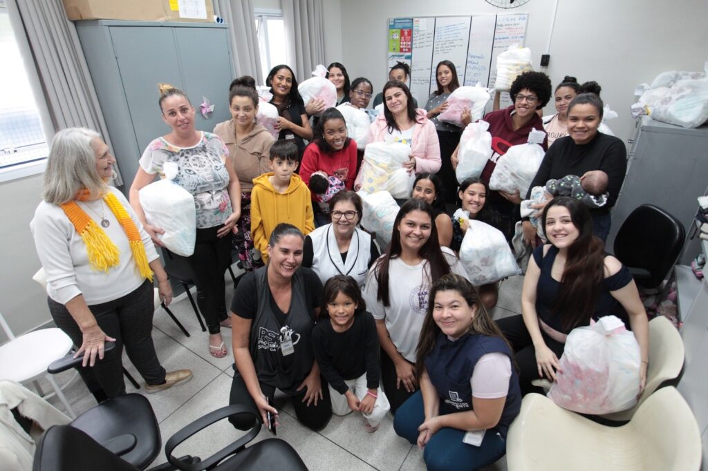 A imagem mostra um grupo de aproximadamente 20 pessoas, principalmente mulheres, reunidas em um ambiente interno que parece ser um escritório ou uma sala comunitária bem iluminada. Muitas delas seguram sacolas plásticas brancas grandes, que aparentam conter doações, possivelmente fraldas ou roupas de bebê. Algumas mulheres estão segurando bebês nos braços, o que sugere que o grupo pode estar envolvido em uma ação de apoio a mães ou famílias com crianças pequenas. No fundo, há um quadro branco com anotações em português, mencionando um comunicado sobre férias em 2026, uma reunião online marcada para 05/04/26 às 10:00, e o nome "Programa Saúde da Gente" e "Mania de Ser". O ambiente é organizado, com cadeiras e armários cinza, e a iluminação é clara e uniforme, provavelmente de luz fluorescente. As pessoas estão posicionadas em pé e agachadas, sorrindo para a câmera, transmitindo um clima de colaboração e solidariedade. Uma mulher usa um jaleco branco, sugerindo que pode ser uma profissional de saúde ou voluntária. A cena sugere um momento de entrega ou distribuição de doações, reforçando um sentimento comunitário e de apoio mútuo.