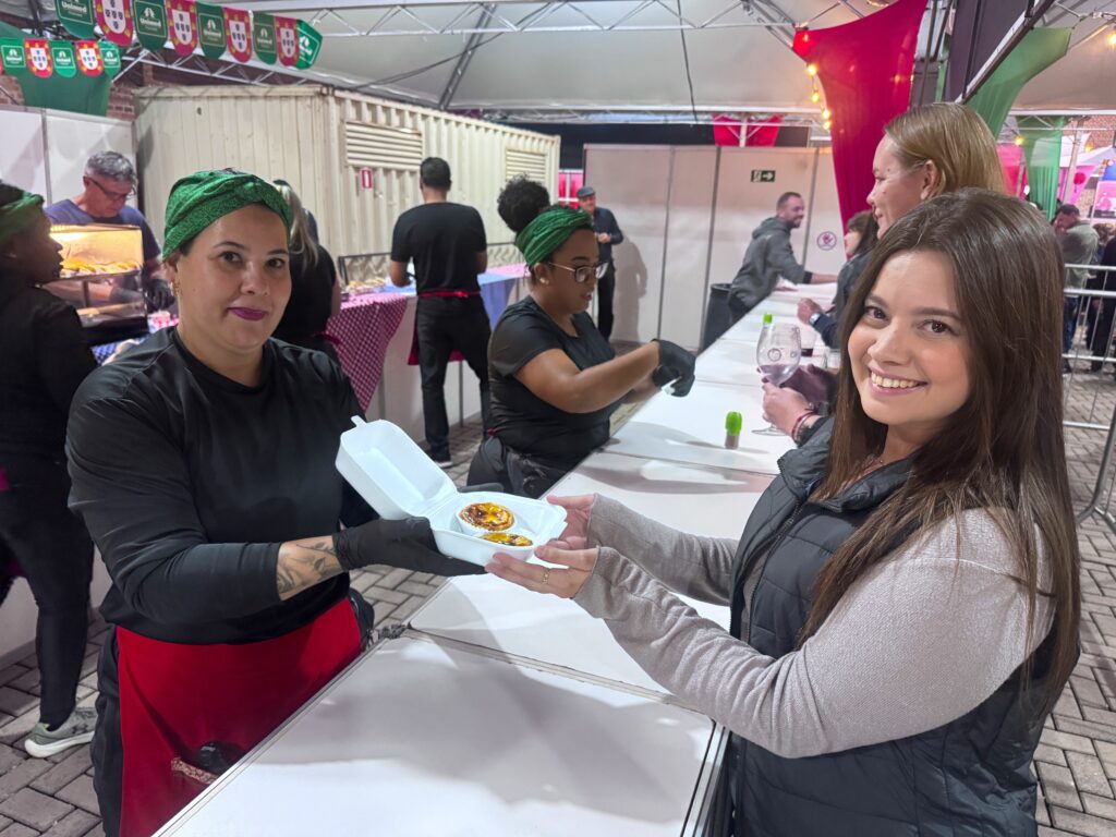 A imagem mostra um cenário ao ar livre, provavelmente em uma feira, festival ou evento gastronômico. No centro da cena, duas mulheres estão interagindo de forma amigável. A mulher à esquerda, que parece ser a vendedora, está usando uma camiseta preta, avental vermelho e um lenço verde na cabeça. Ela está entregando um recipiente branco de isopor com um pastel de nata para a outra mulher, que é a cliente, sorridente, vestindo uma blusa clara com colete preto. A vendedora usa luvas pretas e tem uma tatuagem visível no antebraço. Ao fundo, há outra mulher com um lenço verde na cabeça, óculos e avental vermelho, que parece estar preparando ou organizando alimentos. O local tem mesas brancas alinhadas e a área está decorada com bandeirinhas nas cores verde e vermelha, com a marca "Unimed" visível nas bandeiras. Há outras pessoas ao fundo, algumas observando e outras próximas ao balcão de atendimento. A iluminação indica que o evento ocorre à noite ou em ambiente coberto com luz artificial, criando uma atmosfera acolhedora e festiva. A imagem transmite uma sensação de convivência, troca e celebração em torno da comida.