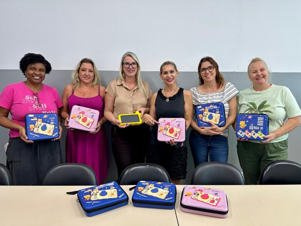 A imagem mostra seis mulheres sorridentes em um ambiente de escritório ou sala de aula, aparentemente apresentando tablets infantis. Elas parecem estar envolvidas em uma ação de entrega ou doação de materiais educacionais. Objetos: A maioria das mulheres segura capas de proteção coloridas (azuis e rosas) com ilustrações, e a mulher ao centro segura um tablet amarelo. Ambiente: Estão posadas atrás de uma mesa com mais estojos dispostos, vestindo roupas casuais. Clima: A cena transmite alegria e um propósito profissional.