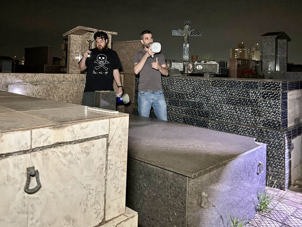 A imagem mostra dois homens em um cemitério durante a noite. O homem à esquerda veste uma camiseta preta com um desenho de caveira e ossos cruzados, acompanhado da palavra "Bu-ri-go". Ele tem barba e segura uma lanterna acesa, que ilumina parcialmente a cena. O homem à direita usa uma camiseta cinza e calça jeans, está segurando um megafone e faz um sinal de positivo com a mão, parecendo estar falando ou interagindo com algo fora do enquadramento. Ao fundo, há várias sepulturas e um grande crucifixo, reforçando o ambiente do cemitério. A iluminação é baixa, típica do período noturno, com luzes artificiais que criam sombras e um clima levemente sombrio. Também é possível ver algumas construções ao fundo, com janelas iluminadas, indicando que o cemitério está próximo a uma área urbana. A imagem transmite uma atmosfera de mistério ou investigação, talvez relacionada a algum evento ou atividade noturna no local.